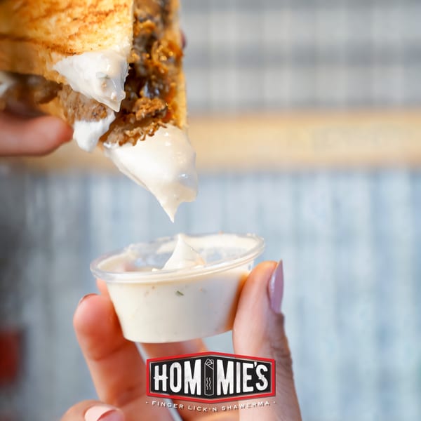A Hommies Shawarma sandwich being dipped into a small cup of white sauce.