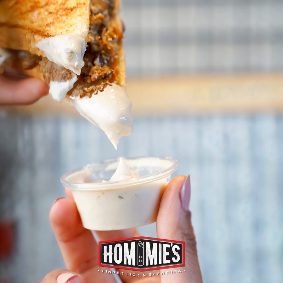 A Hommies Shawarma sandwich being dipped into a small cup of white sauce.