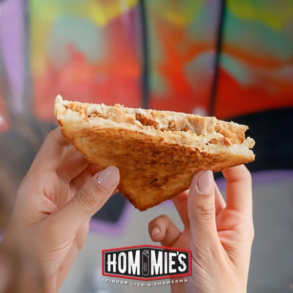 Hands holding a golden-brown, triangular Hommies Shawarma sandwich filled with chicken and white sauce.